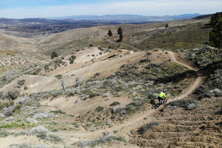 A mountain biker riding down a rocky trail in a hilly, sparsely vegetated landscape, with distant mountains and a small town visible in the background under a clear blue sky.