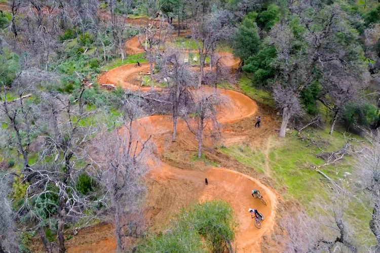 Aerial view of a winding mountain biking trail, surrounded by trees. Two cyclists are riding along the dirt path, which features smooth curves, while a person stands off to the side. The landscape includes a mix of green foliage and bare trees, suggesting a natural environment.