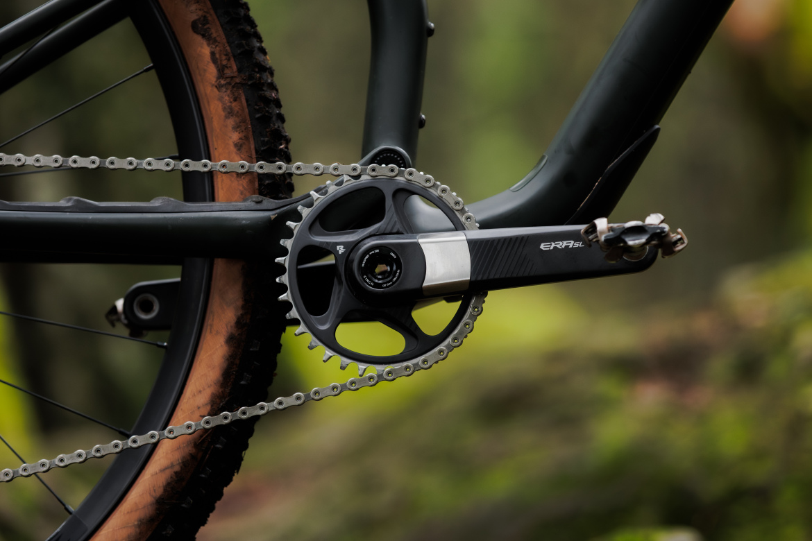 Close-up view of a bike's crankset, showcasing a carbon fiber crank arm, chainring, and chain. The background features blurred greenery, indicating an outdoor setting. The bike has a rugged tire, suggesting it is designed for off-road use.