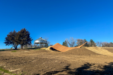 A scenic view of a park featuring a modern, wavy wooden structure and a metal gazebo under a clear blue sky. In the foreground, there is an area of bare earth, suggesting ongoing landscaping or construction. Two evergreen trees are visible next to the gazebo. Carlson Park mountain bike trail.