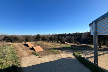 A dirt bike course featuring several brown dirt jumps, set against a clear blue sky and tree line in the background. The foreground includes a gravel path leading to the jumps, with green grass on the edges. Carlson Park mountain bike trail.