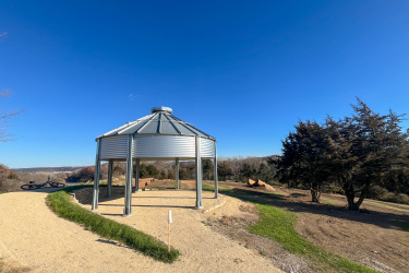 A silver, octagonal gazebo is centrally positioned on a gravel pathway, surrounded by grassy areas and a few trees. The sky is clear blue, and the landscape features rolling hills in the background. To the left, a black bicycle lies on the ground near the path, while earthworks and a dirt jump area are visible farther in the distance. Carlson Park mountain bike trail.