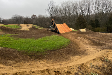 A dirt bike ramp constructed of wood and metal on a grassy dirt track, with winding paths and mounds in the background, under a cloudy sky. Carlson Park mountain bike trail.