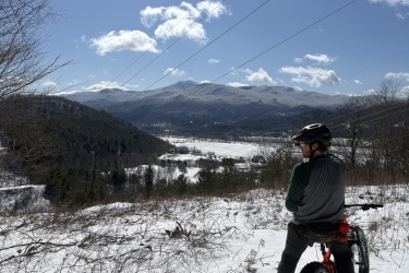 A person sitting on a fat bike, looking out over a snowy landscape with mountains in the distance. The sky is bright blue with some clouds, and the scene is illuminated by sunlight. Trees and brush surround the area, emphasizing the winter setting. Maple Wind Farm mountain bike trail.
