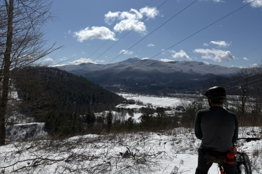 A person on a fat bike sits on a snowy hillside overlooking a valley and mountains in the distance, under a clear blue sky with scattered clouds. Sunlight beams down, illuminating the landscape. Maple Wind Farm mountain bike trail.