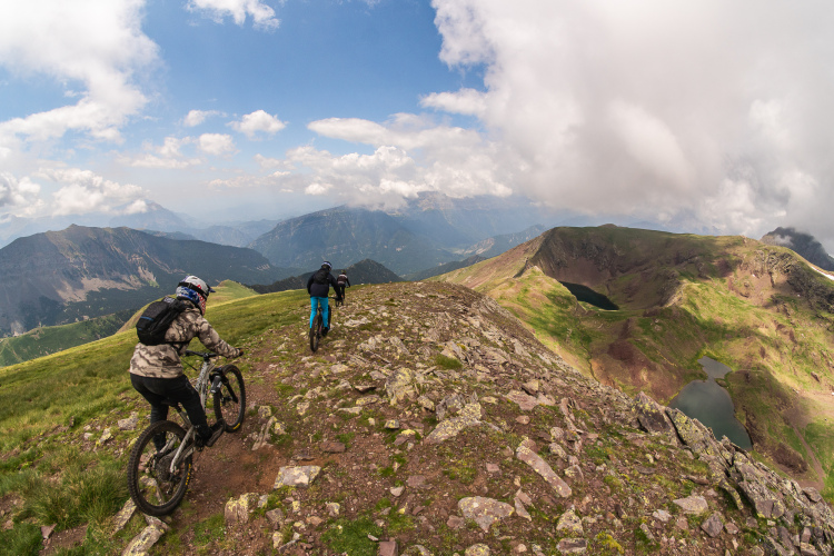 Two mountain bikers navigate a rocky trail on a mountain ridge, surrounded by verdant grass and distant mountains under a partly cloudy sky. The landscape features steep slopes and a serene lake visible in the background, highlighting the natural beauty of the rugged terrain.