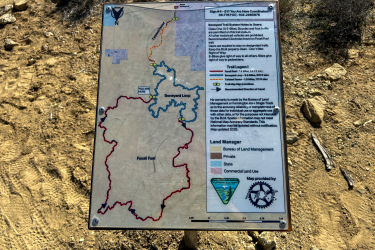 A trail map sign displaying the Boneyard Loop and Fossil Fuel trails, located in a natural outdoor environment. The sign includes directions, trail legends, and notes for users about permissible activities. It also marks the "You Are Here" location and indicates the land manager as the Bureau of Land Management. Boneyard Trails mountain bike trail.