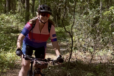 A person riding a mountain bike on a forest trail, wearing a helmet and colorful cycling attire. The surrounding trees are lush with green leaves, and the ground is covered in fallen leaves, indicating a natural outdoor setting. The cyclist is smiling, enjoying the ride in the scenic environment. Twister mountain bike trail.