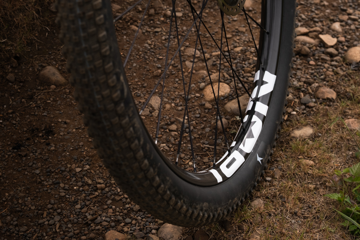 Close-up of a mountain bike tire mounted on a rugged wheel, resting on a gravel and dirt surface. The tire features a prominent brand logo and exhibits a partially worn tread pattern, indicating use in off-road conditions.