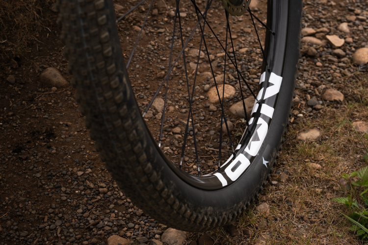 Close-up of a mountain bike tire mounted on a rugged wheel, resting on a gravel and dirt surface. The tire features a prominent brand logo and exhibits a partially worn tread pattern, indicating use in off-road conditions.