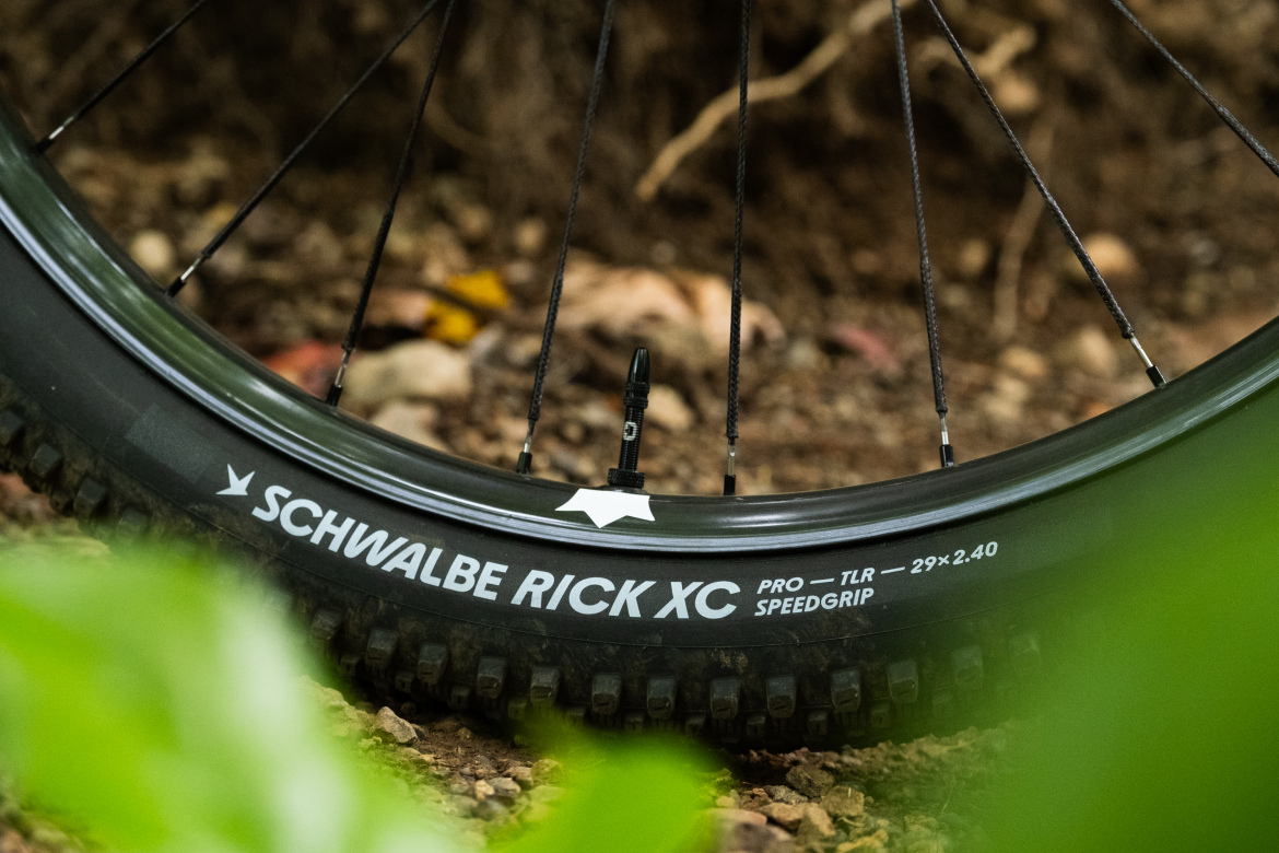 Close-up shot of a bicycle tire labeled "Schwalbe Rick XC Pro - TLR Speedgrip 29x2.40," featuring visible tread patterns and a valve stem, set against a natural background of dirt and foliage.