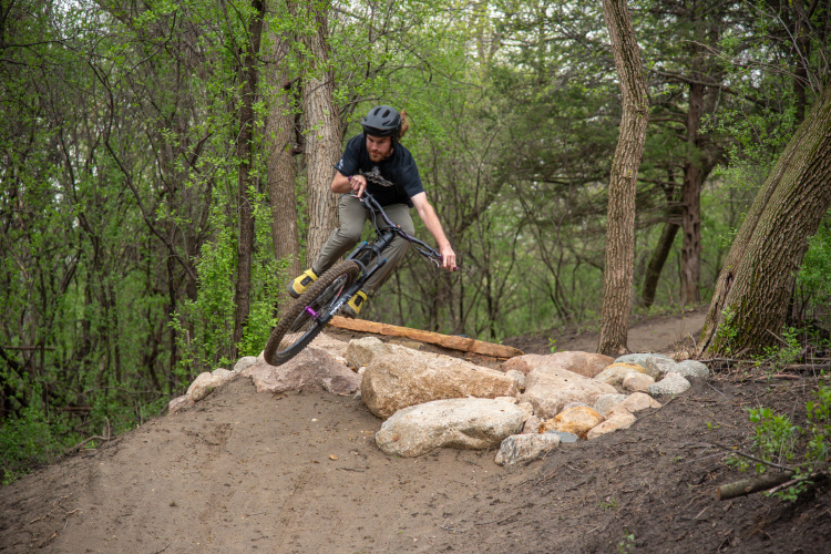 A mountain biker navigating a dirt trail, leaning to the side as they approach a rocky jump in a wooded area. The scene captures the rider mid-action, with trees and greenery in the background.