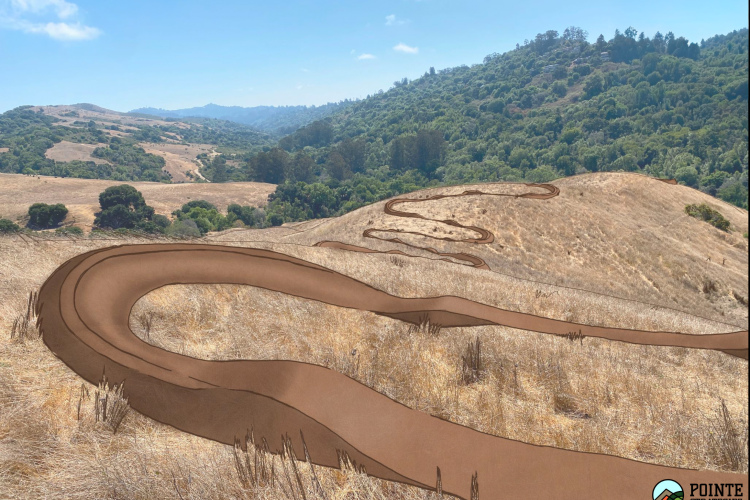 A winding dirt path cuts through a dry, grassy landscape with rolling hills in the background, featuring patches of green vegetation and a clear blue sky. The scene is framed by distant mountains, creating a tranquil outdoor setting.