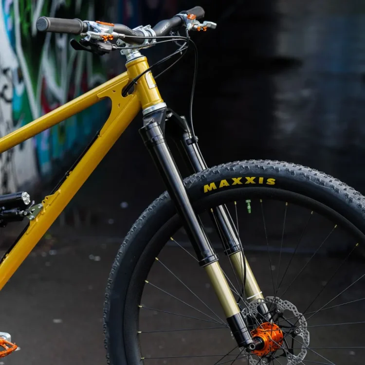 Close-up view of a mountain bike featuring a yellow frame, black suspension forks, and Maxxis tires. The handlebars are equipped with orange accents, and a disc brake is visible on the front wheel. The background includes a blurred, graffiti-covered wall.