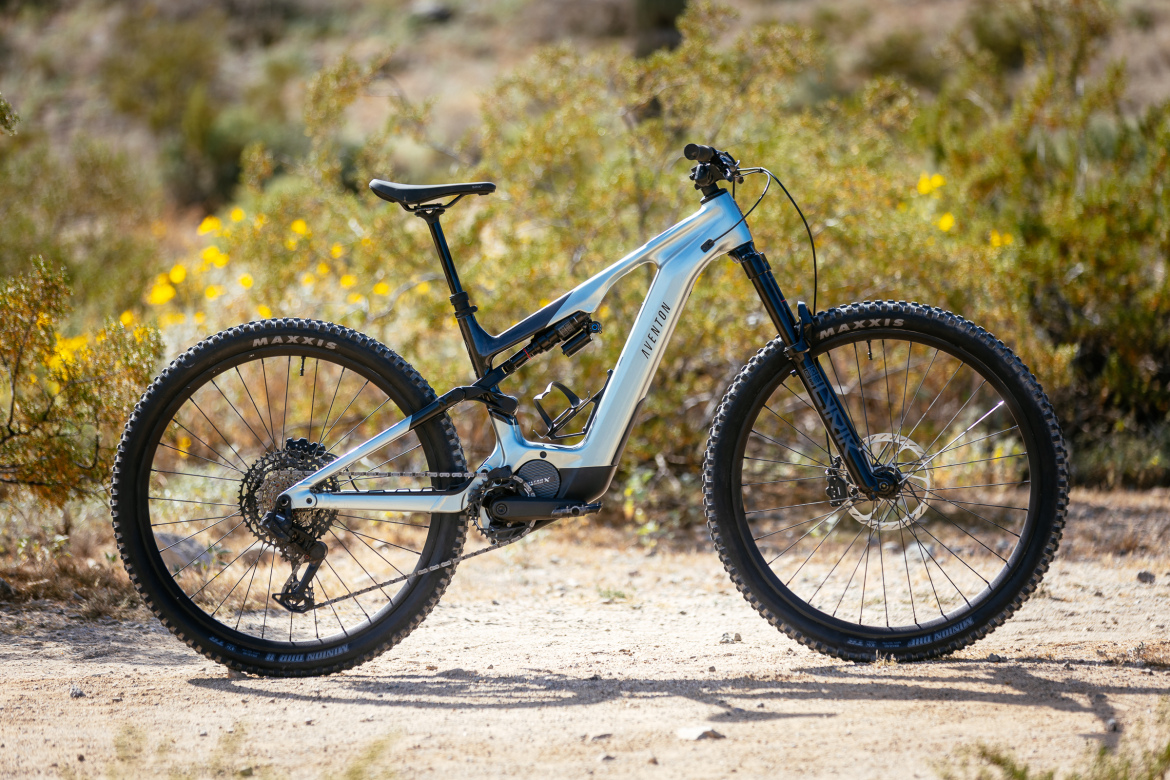 A modern mountain bike with a sleek design, featuring a metallic blue frame, dual suspension, and rugged tires set against a natural background of shrubs and wildflowers. The bike is positioned on a dirt trail, showcasing its suitability for off-road terrain.