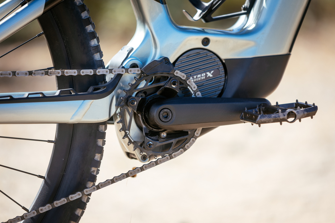 Close-up view of a mountain bike's drivetrain, showcasing the crankset, chain, and rear tire. The bike features a silver frame and a black chainring, with the chain visibly engaged on the gears. The background is blurred, emphasizing the bike's mechanical components.