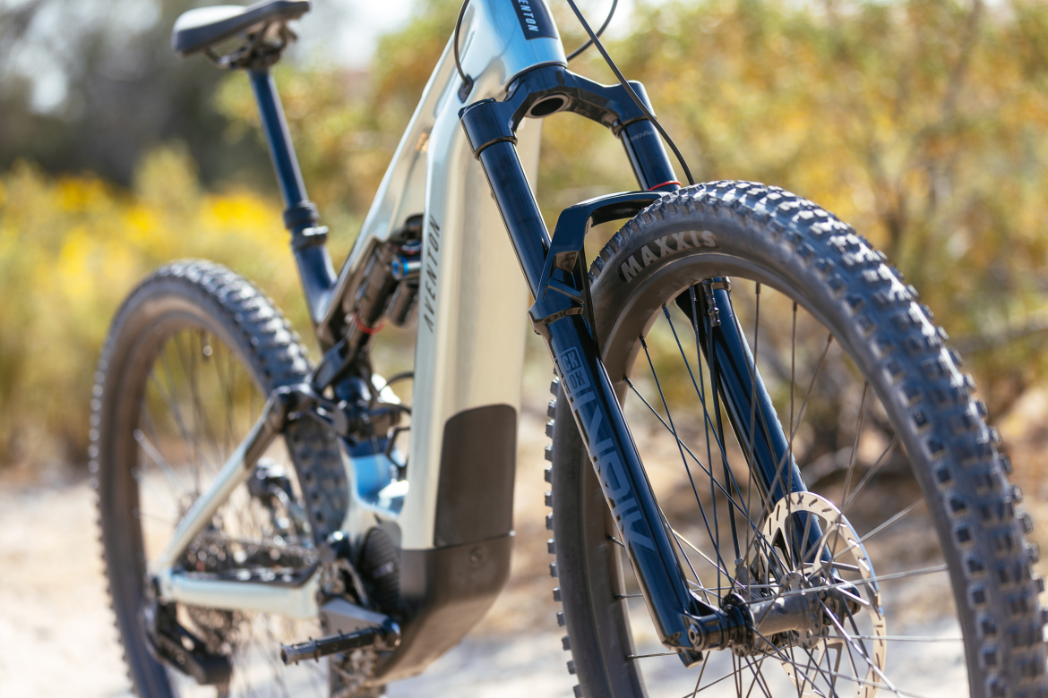 Close-up view of a mountain bike featuring a suspension fork and thick, knobby tires. The bike's frame is light blue with branding visible, and it is set against a blurred background of greenery and wildflowers, suggesting an outdoor setting.