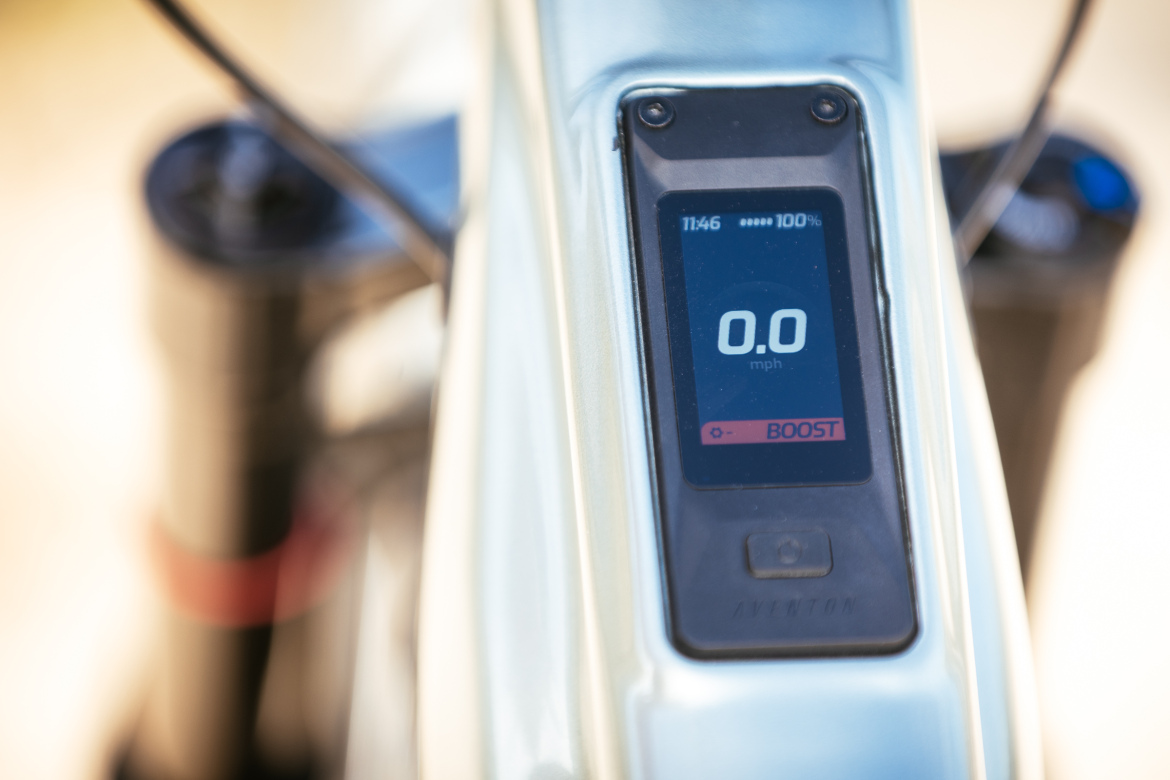 Close-up of an electric bike's display screen showing a speed of 0.0 mph, a battery level of 100%, and the time as 11:46. The screen is set against the bike’s frame, with a boost button visible.