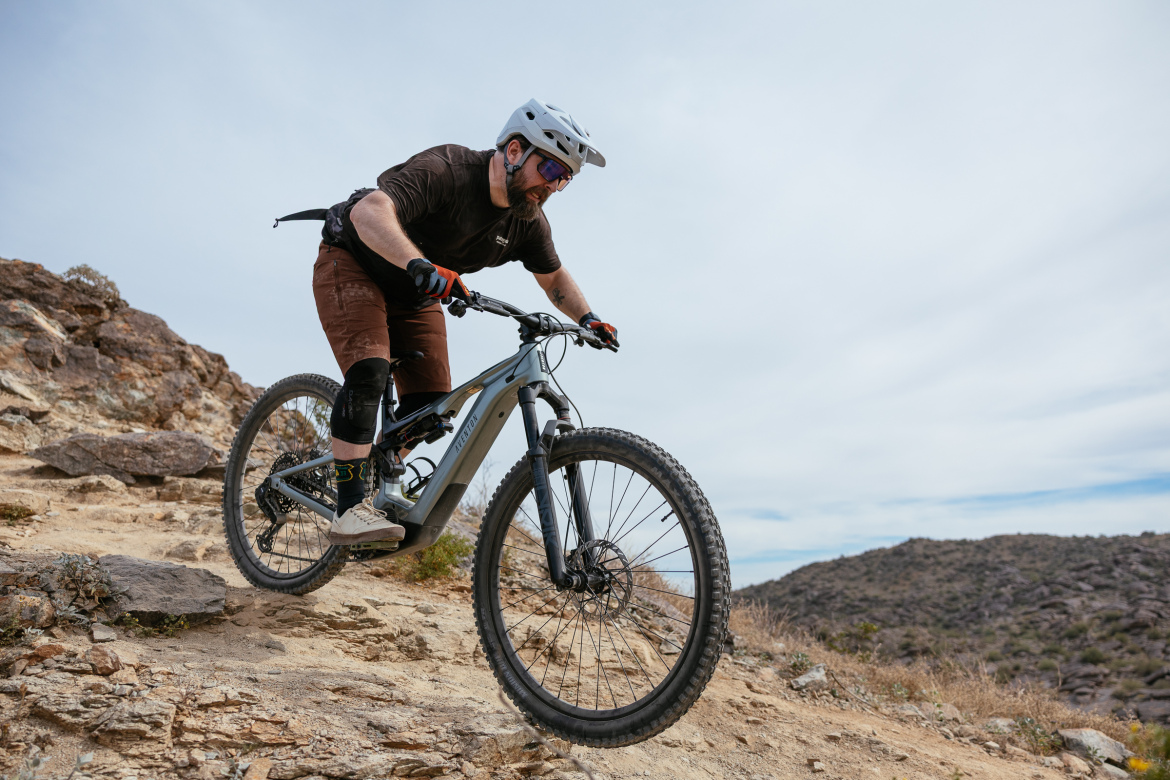 A person riding an electric mountain bike down a rocky trail, focused on maintaining balance. The rider is wearing a helmet, gloves, and protective gear. In the background, there are hills and a cloudy sky.