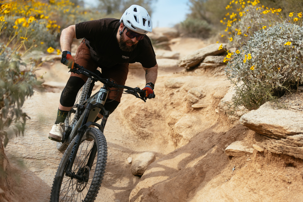 A mountain biker riding on a dirt trail surrounded by desert flora, including yellow wildflowers. The biker is wearing a white helmet, sunglasses, and protective gear, navigating a rocky section of the path with focus and skill. The landscape features a mix of sandy soil and rocky terrain under a clear blue sky.