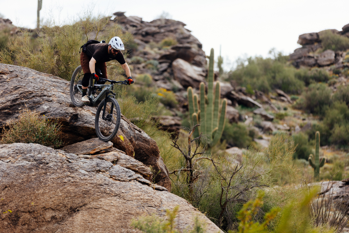 A mountain biker skillfully navigates a rocky terrain, perched on a large boulder with cacti and desert vegetation in the background. The rider wears a helmet and protective gear, displaying focus and balance as they tackle the challenging landscape.