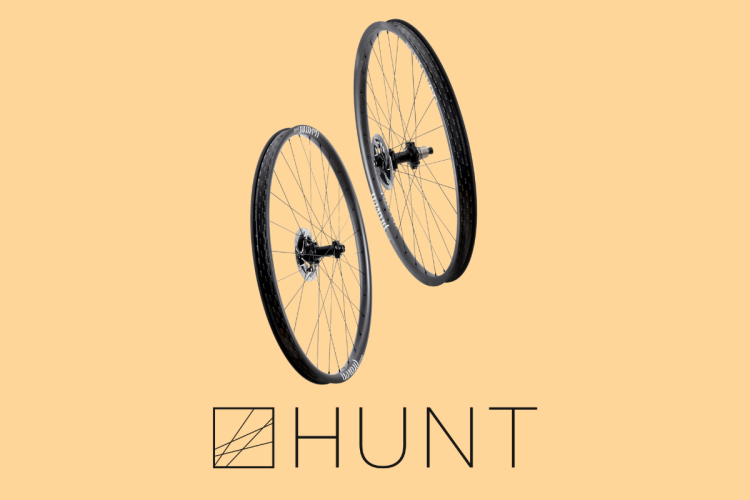 Two bicycle wheels are displayed against a light orange background, with the word "HUNT" prominently featured below them. The wheels are black, showcasing their spokes and hubs, and are positioned to create a dynamic composition.