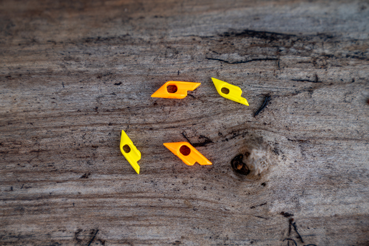 Four small, colorful plastic pieces in yellow and orange are scattered on a wooden surface. The pieces have angular shapes with circular holes, resembling components that could be used in a game or tool. The rustic texture of the wood contrasts with the bright colors of the plastic.