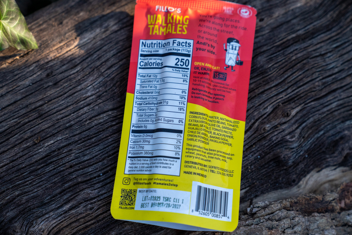 A colorful package of Filo's Walking Tamales resting on a wooden surface, showcasing the nutrition facts and ingredients. The front of the package includes the product name, serving size, and calorie information. The ingredients list and preparation instructions are printed on the back, along with a QR code and best-by date.