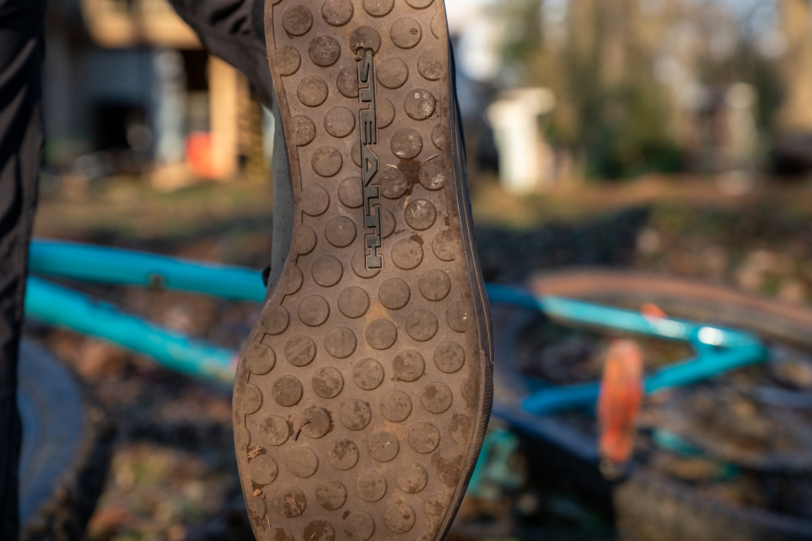 Close-up of a muddy shoe sole with a dotted tread pattern, held up in the foreground, with a teal bicycle partially visible in the background.