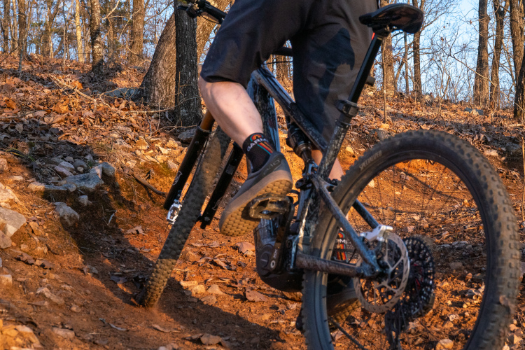 A mountain biker navigating a rocky trail in a forested area, focusing on the rear wheel and pedals of the bike. The ground is covered with orange dirt and scattered leaves, and sunlight filters through the trees in the background.