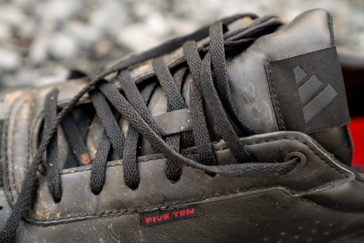 Close-up of a black sneaker with a textured leather upper and black laces. The shoe features a visible logo on the side and a small red tag that says "FIVE TEN." The background shows a blurred, rocky surface indicating an outdoor setting.