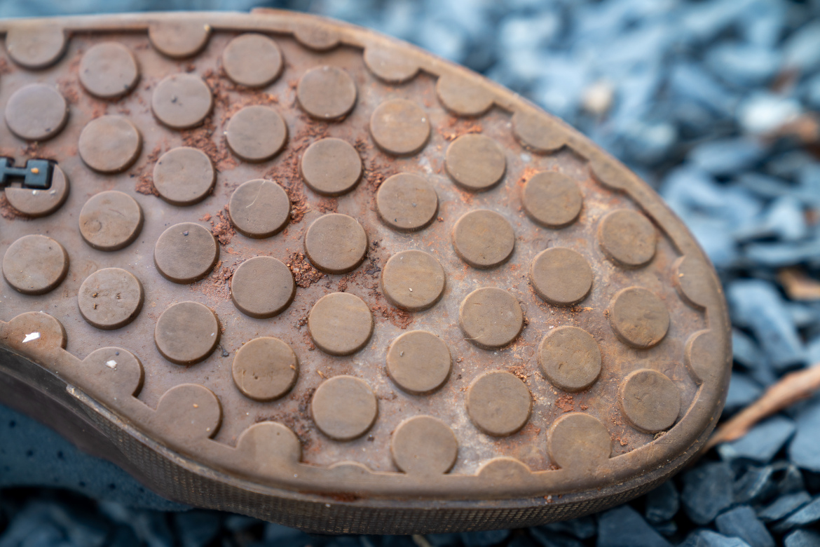 Close-up image of the sole of a worn shoe, showing a pattern of circular treads and dirt particles. The background features small, dark gravel stones.