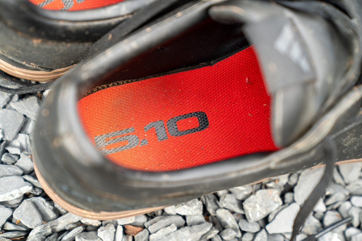 A close-up view of the interior of a black athletic shoe, showing a bright red insole with the text "5.10" printed on it. The shoe is placed on a surface of small gray stones, indicating outdoor use.