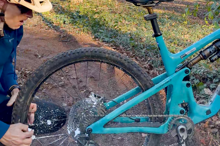A cyclist kneeling beside a mountain bike, inspecting the rear tire, which is covered in white foam likely from a sealant for puncture repair. The background features a dirt path surrounded by greenery, indicating an outdoor biking environment. The cyclist is wearing a helmet and a blue shirt.