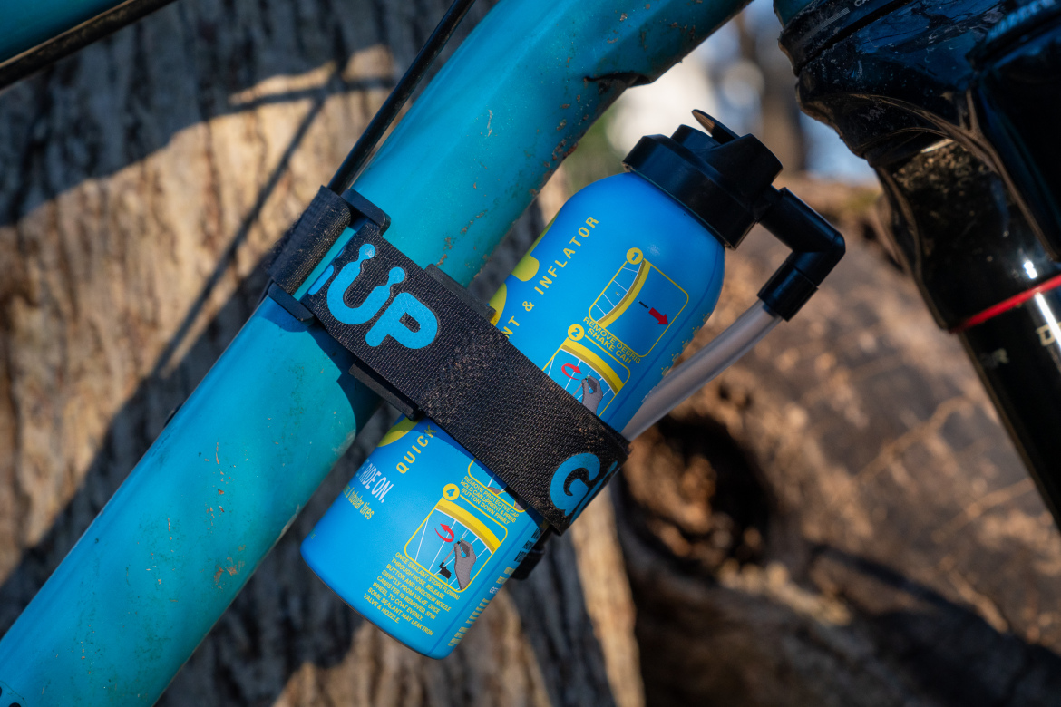 A blue bike-mounted inflator bottle secured with a black strap, positioned beside a teal bicycle frame. The bottle features instructional graphics for quick inflation and deflation. In the background, a blurred natural setting with tree bark is visible.