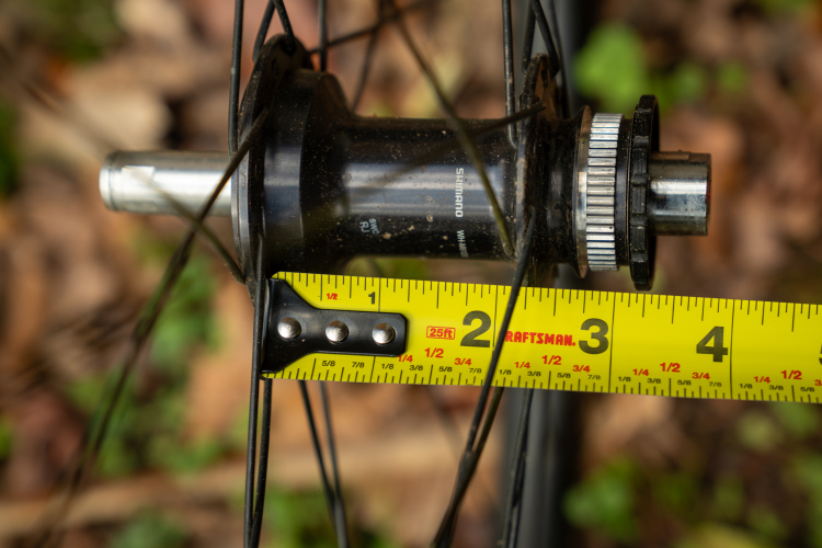 Close-up image of a bicycle hub with a measuring tape showing a measurement close to 2.5 inches. The hub is black and partially dirty, indicating it may be used or older. The background consists of blurred natural elements, likely leaves and grass, suggesting an outdoor setting.