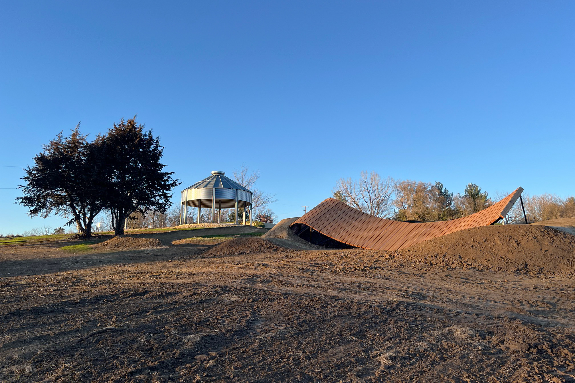 An empty outdoor park scene featuring a circular gazebo with metal roof and supportive columns, situated on a grassy hill beside two large trees. In the foreground, there is an abstract, curved structure resembling a ramp or artistic installation, surrounded by dirt and unlandscaped ground under a clear blue sky. Carlson Park mountain bike trail.