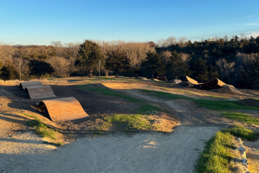 A dirt bike park featuring multiple curved wooden ramps and mounds, situated on a hillside with sparse trees in the background. The sky is clear and blue, casting soft sunlight over the terrain, which includes both grassy and dirt paths. Carlson Park mountain bike trail.