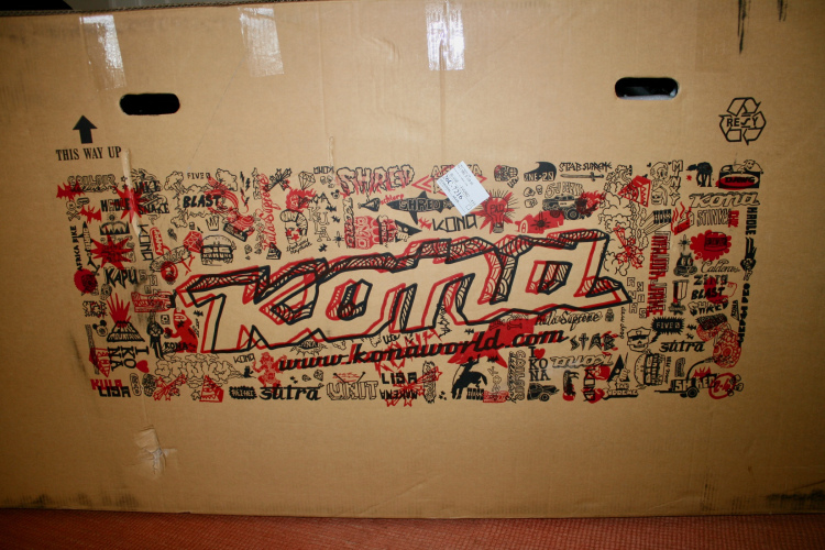 Cardboard box featuring a vibrant design with the word "Kona" prominently displayed in red, surrounded by various doodles and icons related to biking and outdoor activities. The box indicates "THIS WAY UP" and includes a small shipping label.