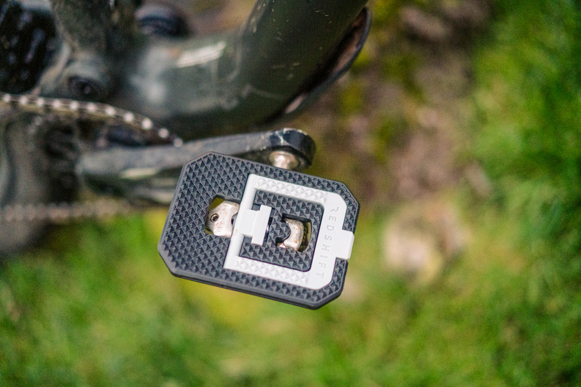 A close-up image of a bike pedal attachment, featuring a black and silver design with a textured surface. The attachment is connected to a bicycle's crank arm, set against a blurred background of green grass.