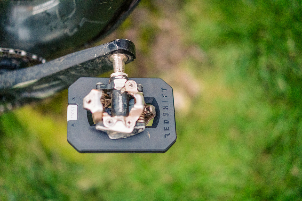 A close-up image of a bicycle pedal featuring a black platform design with a silver attachment point. The pedal is positioned above a grassy area, showcasing some dirt marks, indicating usage. The brand name "REDSHIFT" is visible on the side of the pedal.
