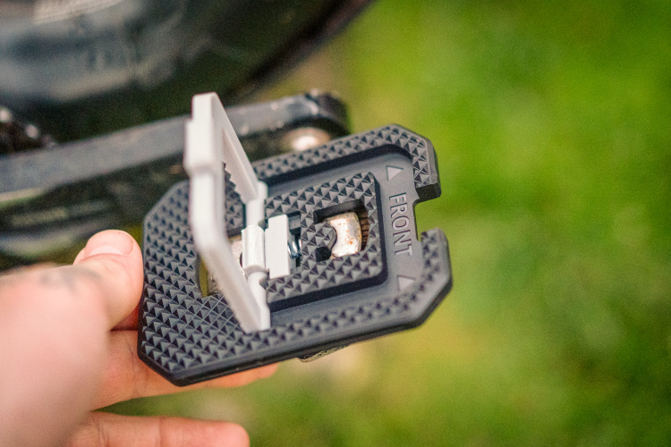 A close-up image of a hand holding a black and gray camera quick release plate, featuring a textured design and the word "FRONT" embossed on one side. The background shows a blurred green outdoor environment.