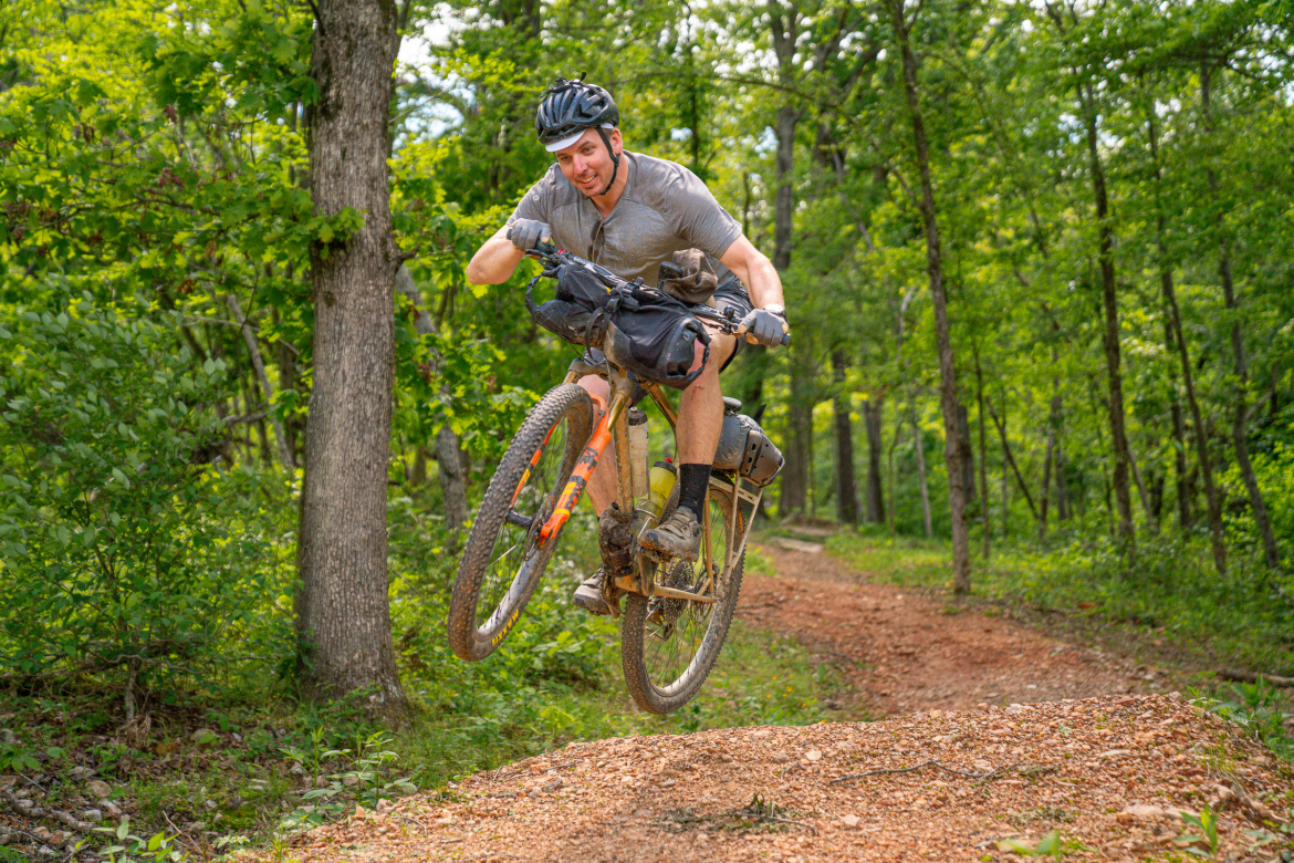 A mountain biker performing a jump on a dirt trail surrounded by trees, smiling as he propels off the ground with his bike. The biker is wearing a helmet, gloves, and casual clothing, and the terrain is a mix of gravel and natural foliage.