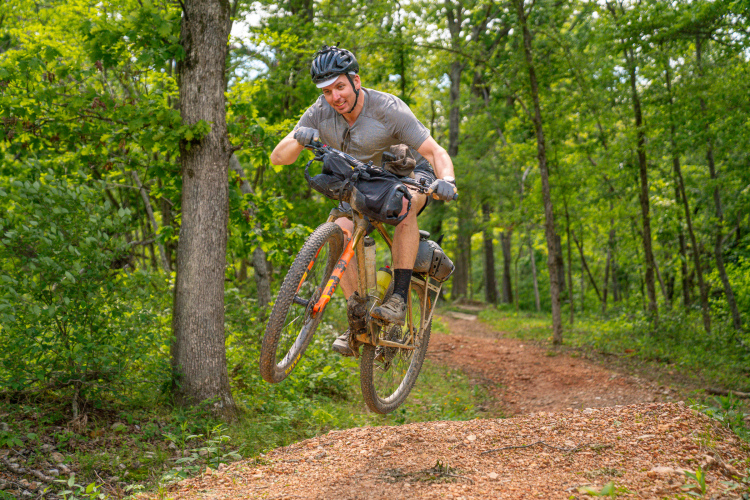 A mountain biker performing a jump on a dirt trail surrounded by trees, smiling as he propels off the ground with his bike. The biker is wearing a helmet, gloves, and casual clothing, and the terrain is a mix of gravel and natural foliage.