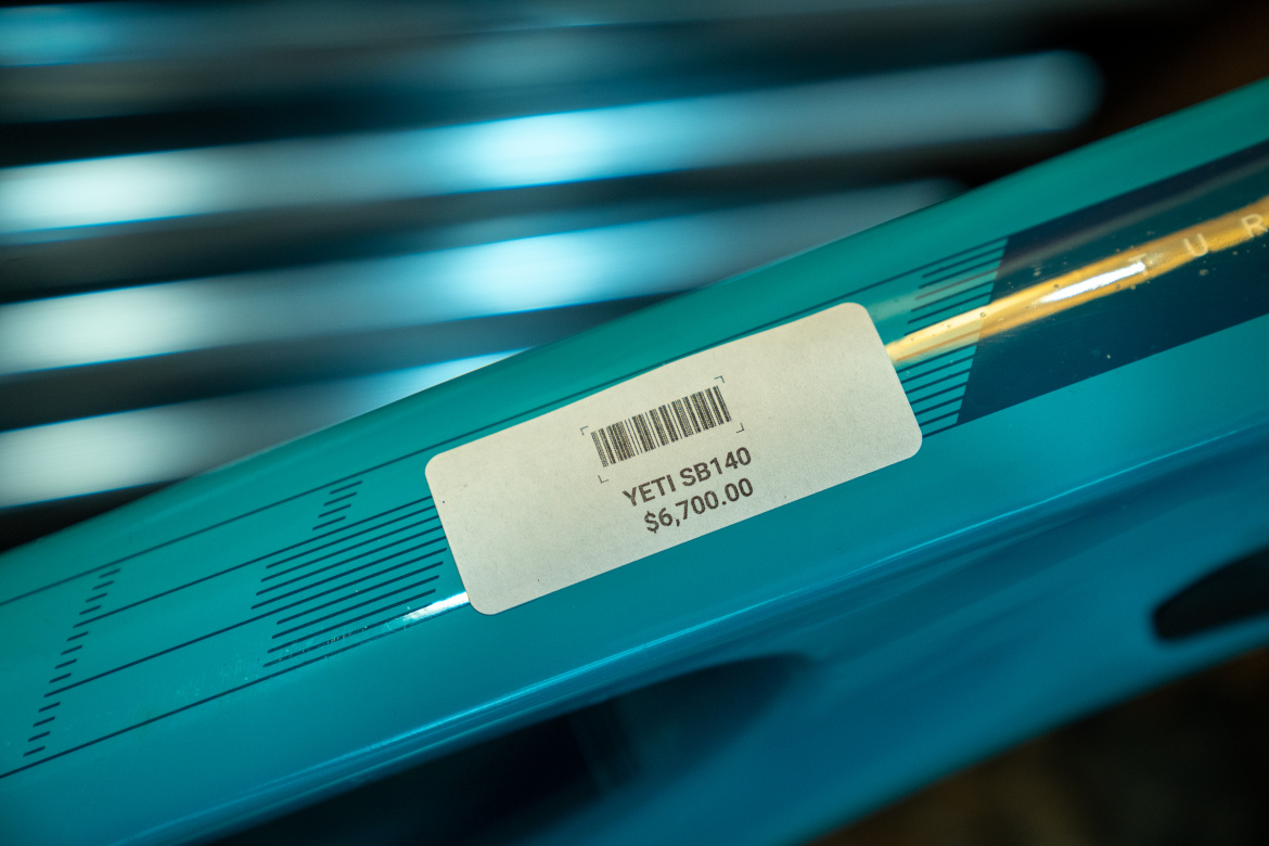 Close-up image of a blue mountain bike frame featuring a price tag that reads "YETI SB140" and a cost of "$6,700.00." The frame has graphic designs and is partially blurred in the background, emphasizing the price tag.