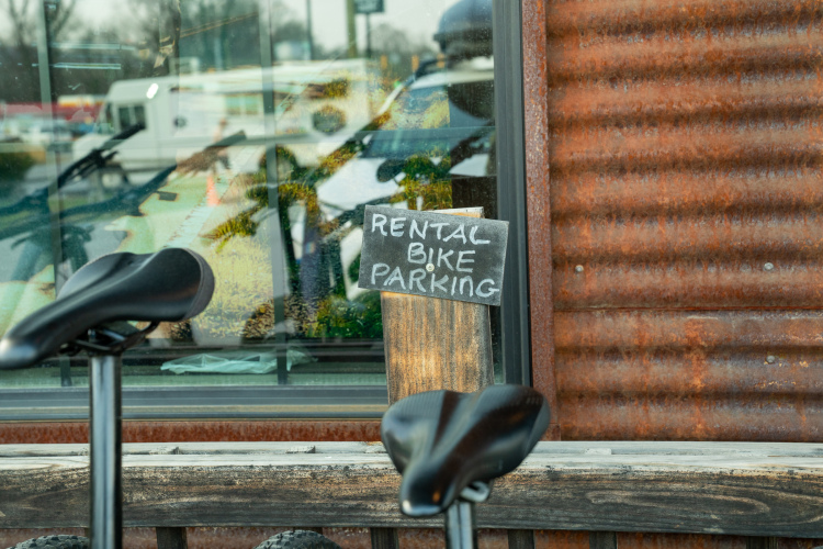 A close-up of a wooden sign that reads "RENTAL BIKE PARKING," placed in front of a window with reflections of bicycles and a busy street. In the foreground, part of a bicycle seat is visible, along with a rustic wooden bench. The background features a textured, rusty metal wall.