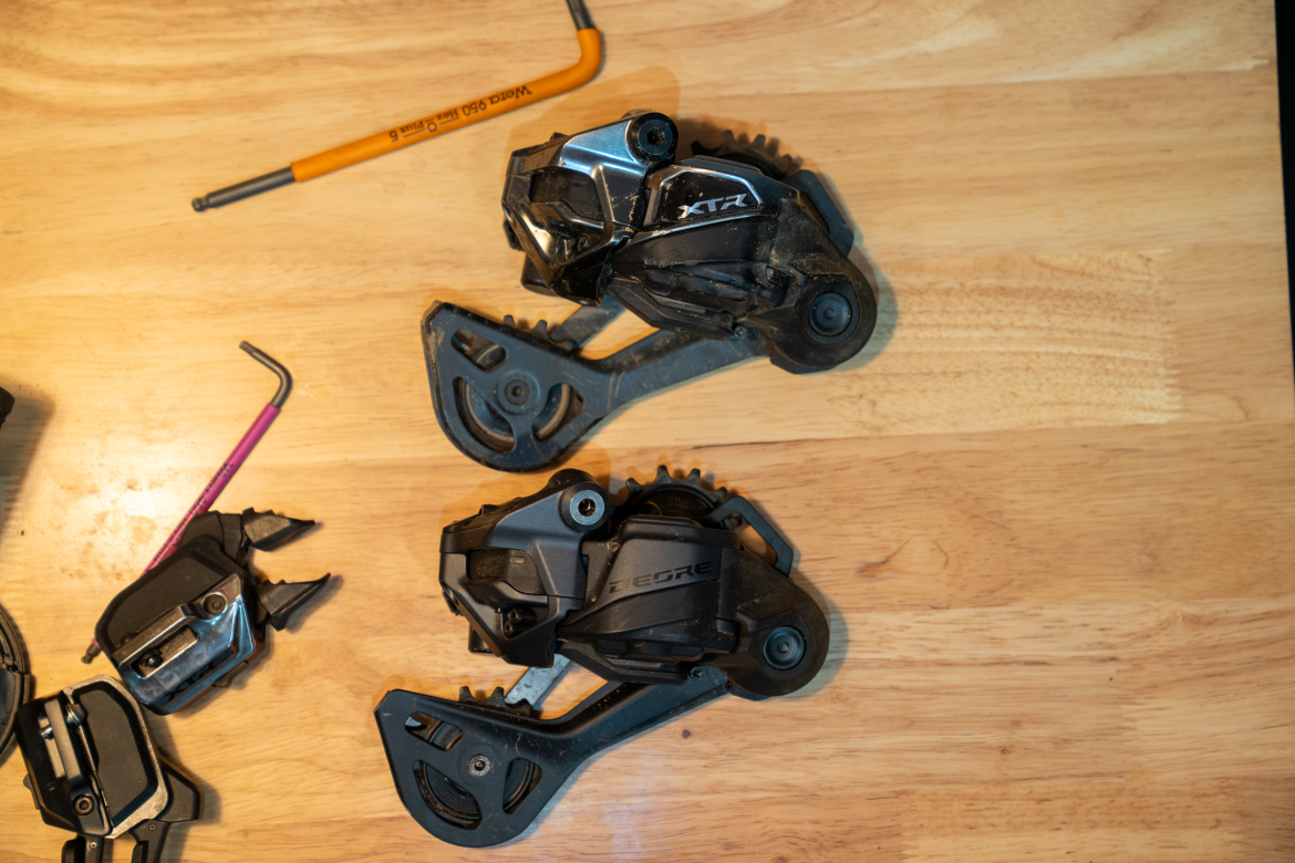 Two bicycle derailleurs are displayed on a wooden surface. The upper derailleur features the "XTR" branding, while the lower device has "GRX" marked on it. Nearby, a pink hex key and a yellow pencil are visible, suggesting a maintenance or repair task.