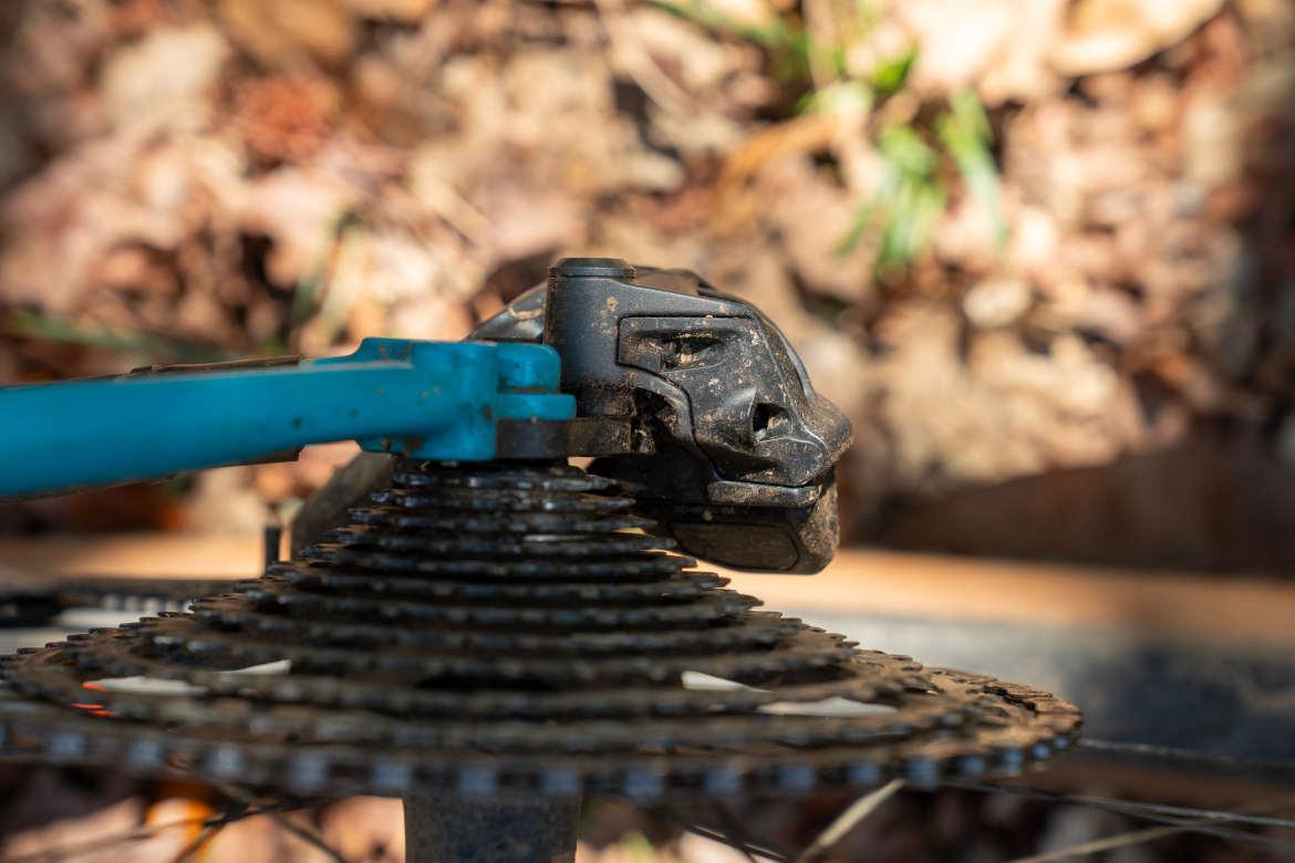 Close-up of a bicycle derailleur positioned near a cassette, with dirt and debris visible on the components, set against a blurred background of foliage and leaves.