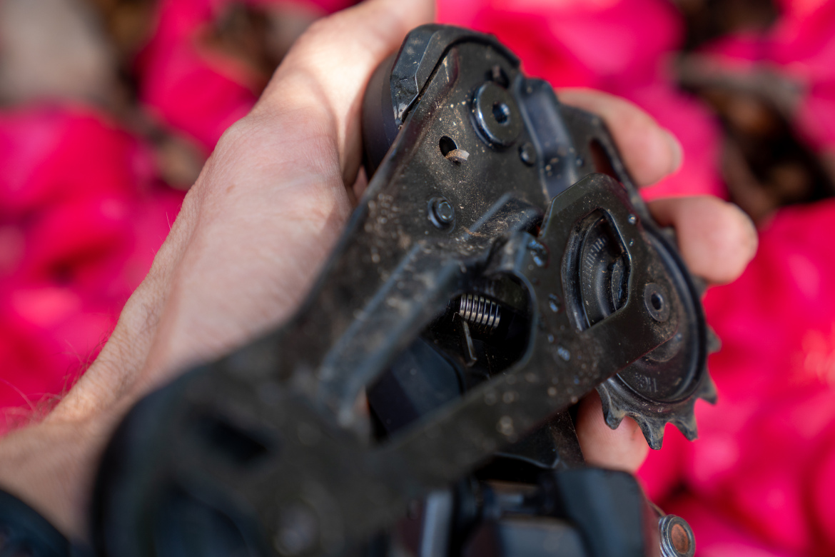 A close-up image of a person's hand holding a black bicycle derailleur, showcasing its mechanical components. The background has a soft focus of bright pink flowers, contrasting with the dark metal of the derailleur.