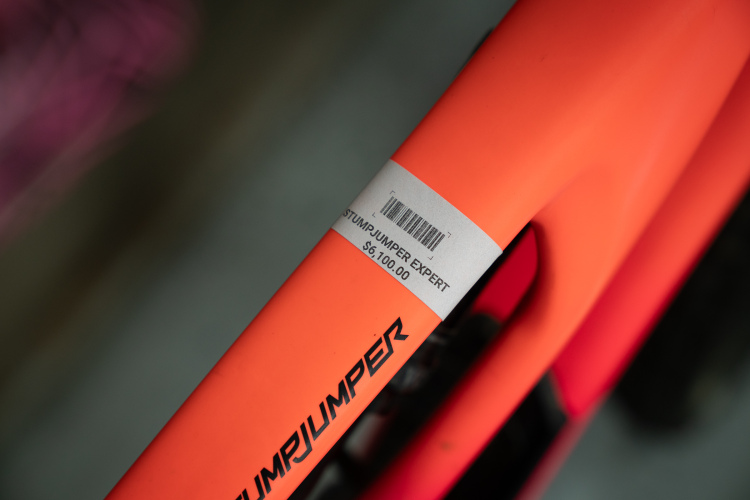 Close-up view of a bright orange bicycle frame labeled "STUMPJUMPER EXPERT" with a price tag indicating $6,100. The background is blurred, focusing on the frame and the tag details.